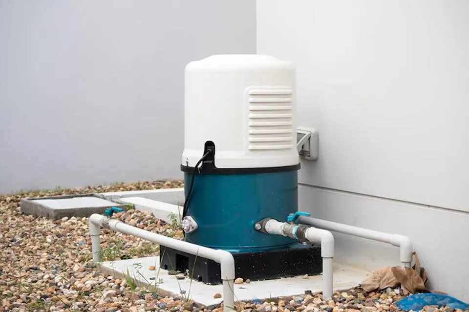  A residential water pump system installed outdoors on a gravel surface next to a white building. The pump is enclosed in a blue and white casing and connected to several white PVC pipes. The setup appears to be part of a home water supply or irrigation system.