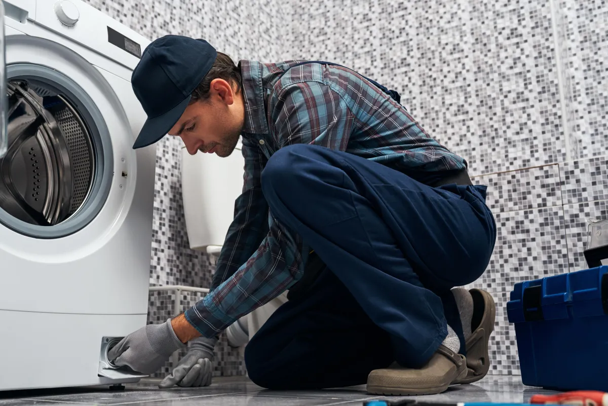 A maintenance worker wearing a plaid shirt, gloves, and a cap kneels on the floor while repairing a front-loading washing machine. The setting is a tiled laundry room, with a blue tool box and various tools visible nearby.