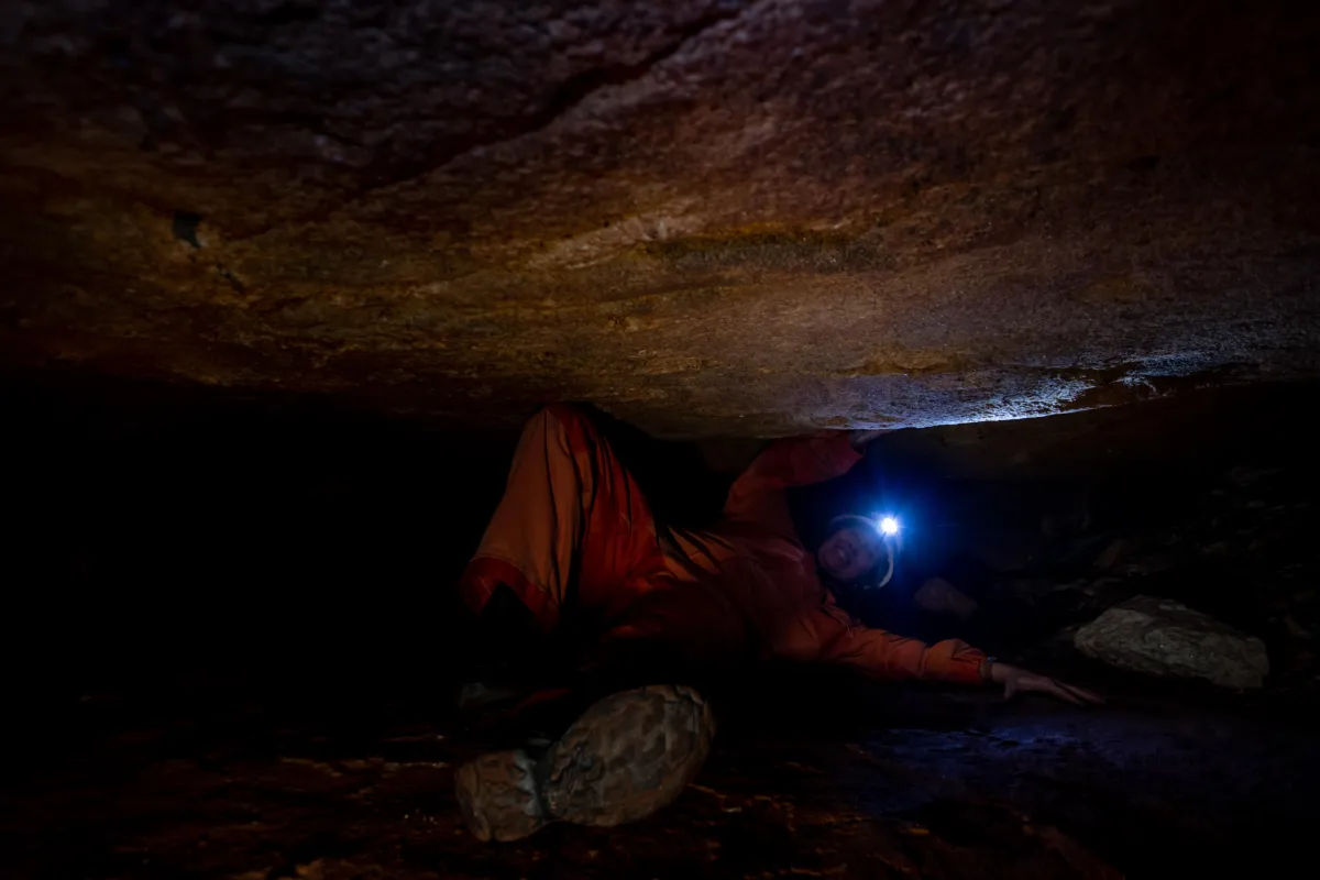 A person wearing an orange jumpsuit crawls through a narrow cave passage while holding a flashlight. The dim lighting and tight space emphasize the confined and rugged environment, typical of spelunking or cave exploration.