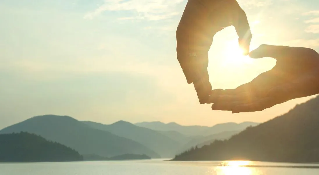 Two hands form a heart around the sun as it hangs low over a mountain landscape.