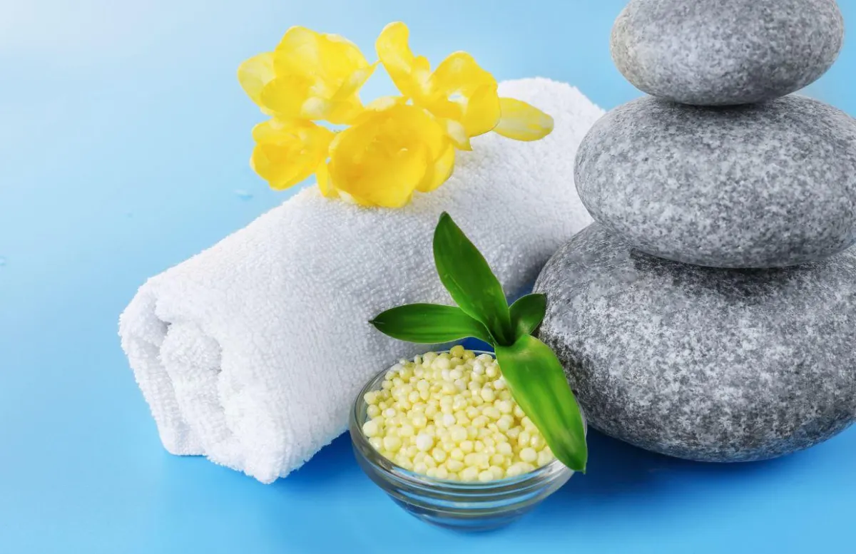 A cairn of massage stones, a rolled blanket topped with yellow orchids, and a bowl of yellow kernels on a blue background.