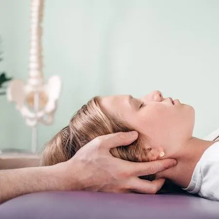 A woman's head on a massage table is held by two hands. Her eyes are closed. A spinal anatomy sculpture is in the background.