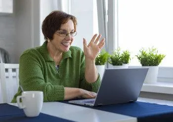 A middle aged woman wearing green smiles and waves to someone on a laptop screen