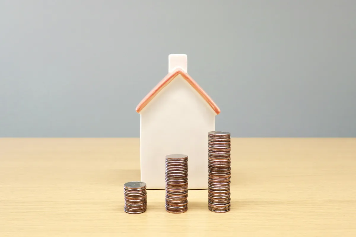 A small wooden model house next to stacks of coins on a desk, representing property taxes and home costs for Michigan buyers.