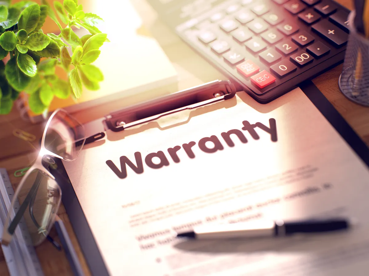 Clipboard with the word Warranty beside a calculator, eyeglasses and a green plant on a desk