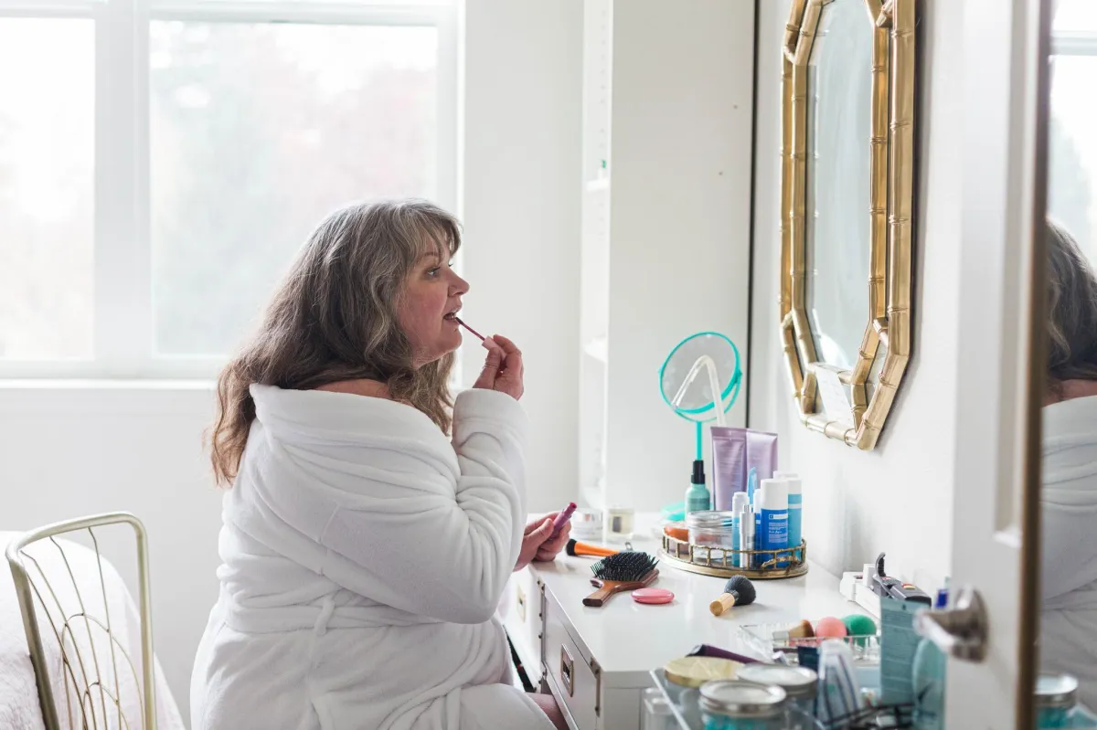 white woman doing makeup in robe I front o gold mirror teal mirror bright window