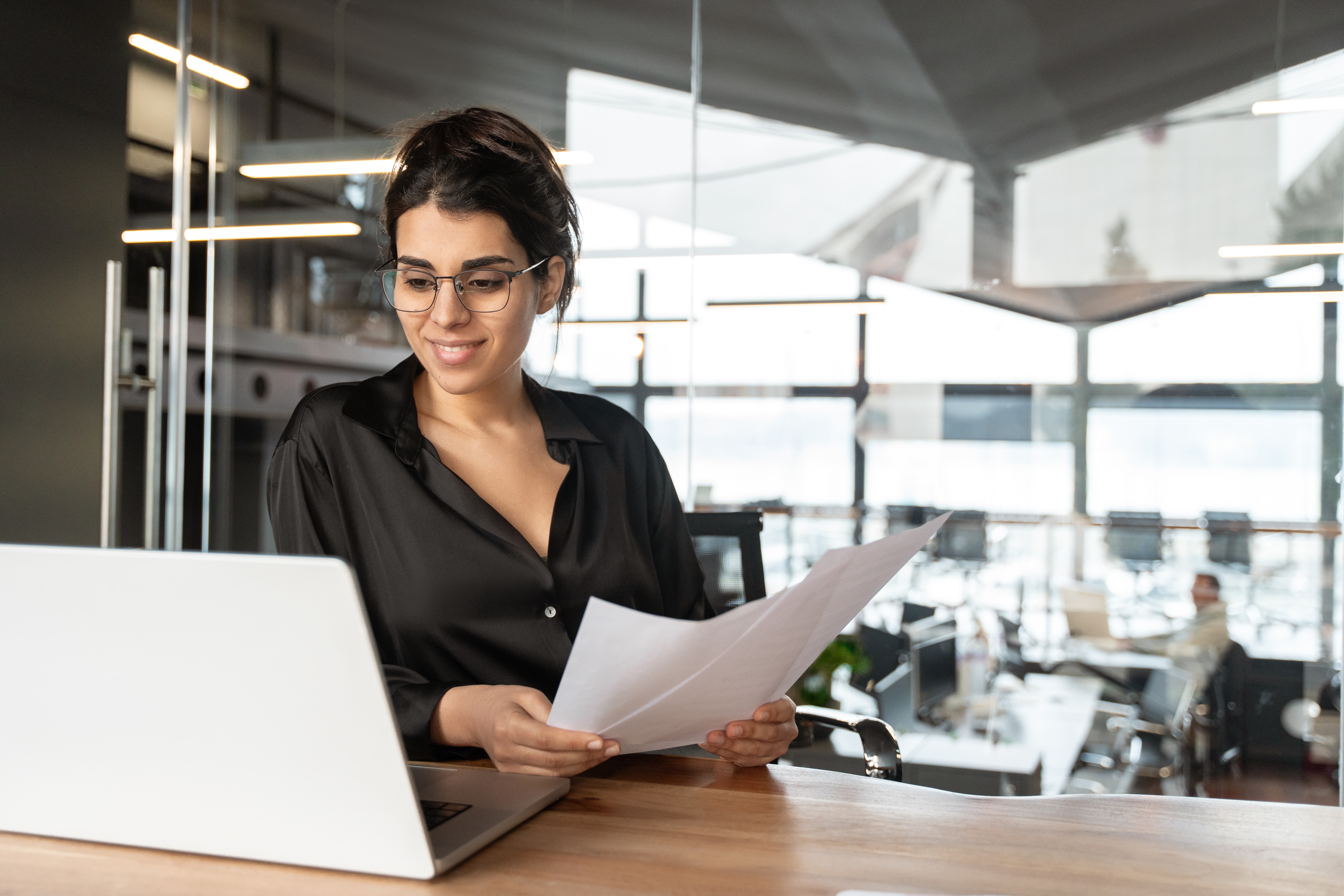 Woman in 30s working at desk holding papers with light in windows.