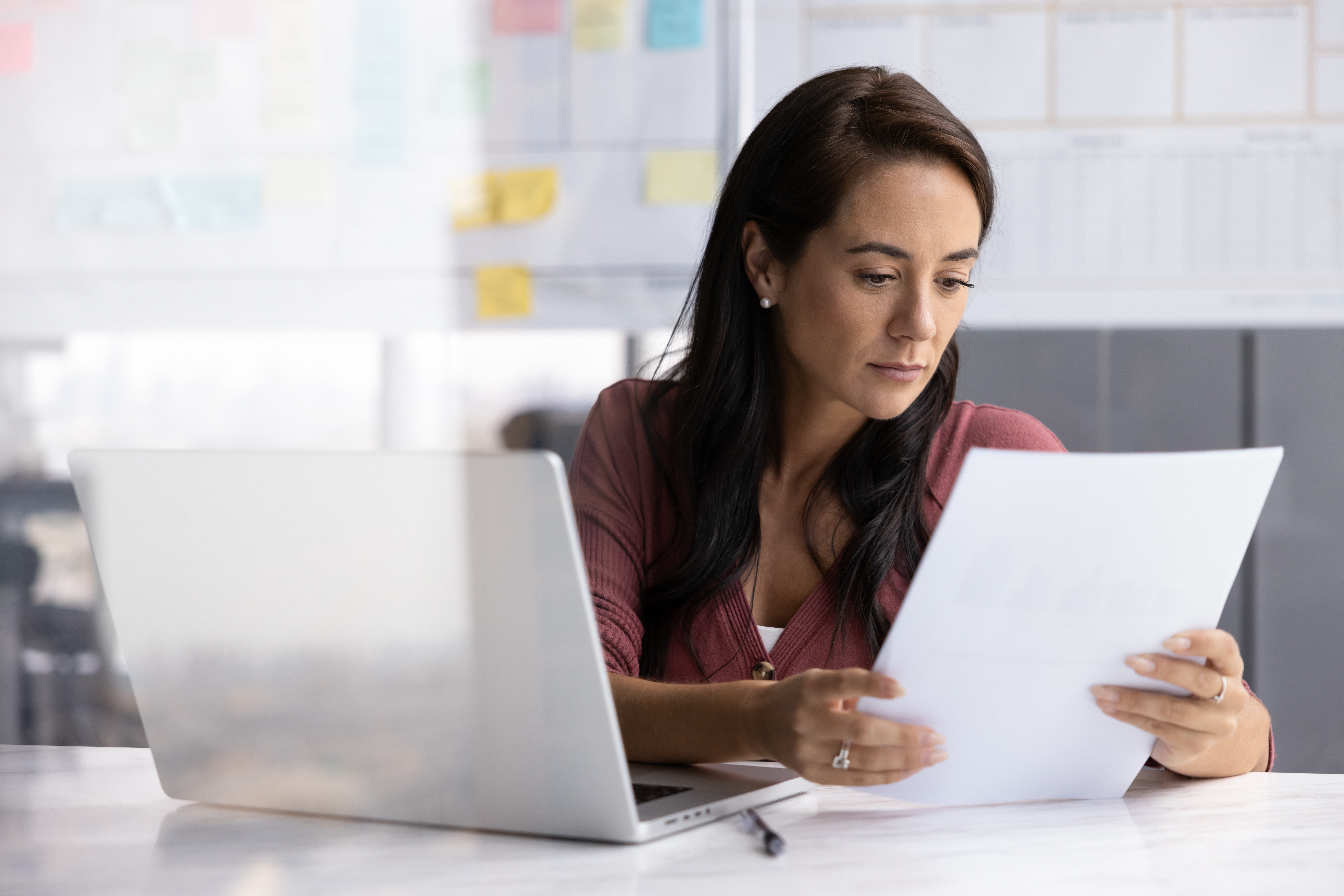 Woman in 40-50s working at desk holding papers with light reflecting strategy board.