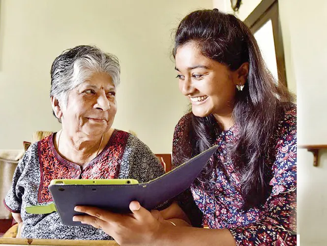 A smiling caregiver sits with an older woman at home, looking at a tablet together