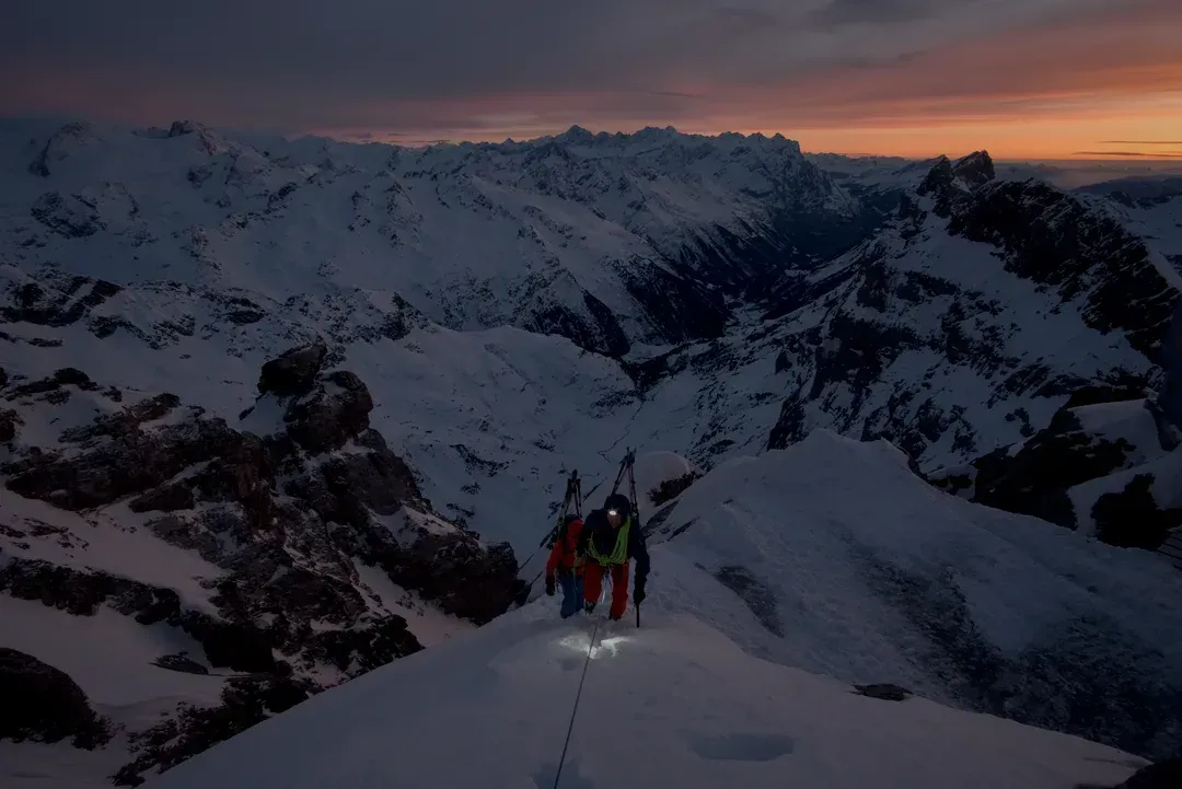 Zwei Alpinisten steigen im Schein der Stirnlampe beim Sonnenuntergang einen Grat hoch.