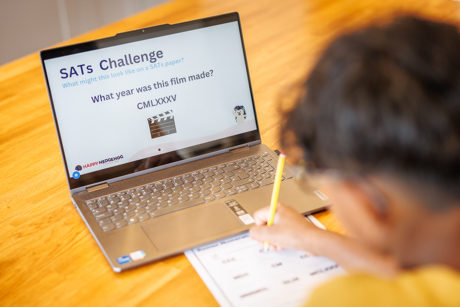 A child working through a SATs Challenge question at a laptop with pencil and paper.