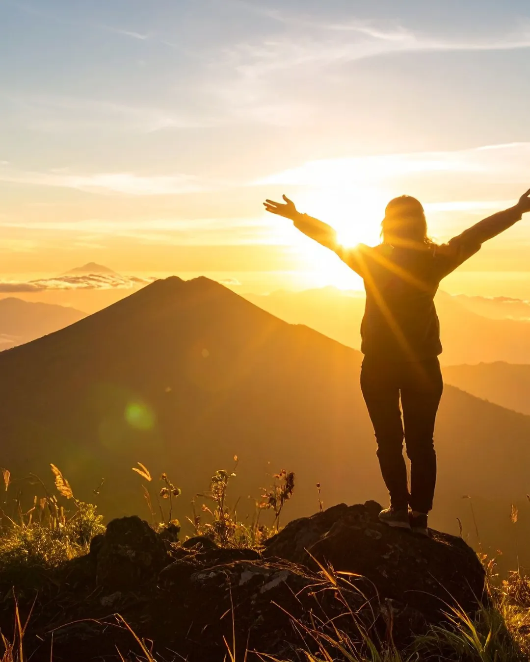 A person stands at a mountain summit with arms raised at sunset, celebrating long-term weight loss success