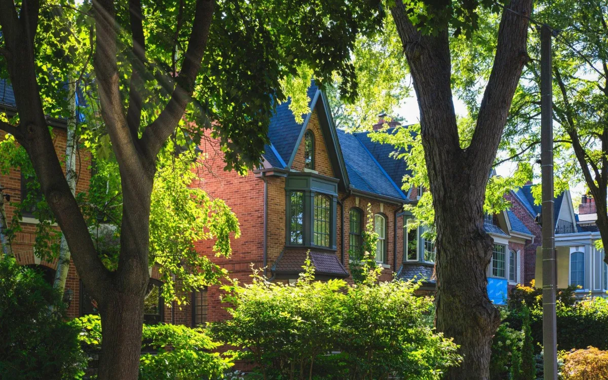 Sun-dappled tree-lined street with a Victorian brick home, bay window, and mature summer canopy, the quiet, established character of Rosedale and Forest Hill, Toronto.