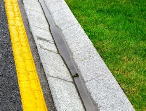 A close-up view of a road curb with a yellow line, a narrow strip of water in the gutter, and a patch of green grass alongside the curb.