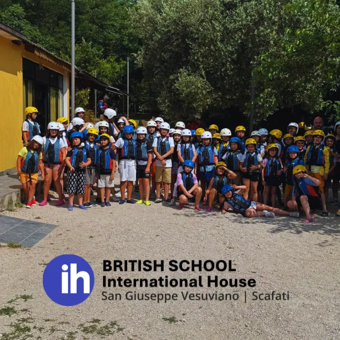 Una foto di gruppo all'aperto di studenti della British School International House San Giuseppe Vesuviano | Scafati, che indossano caschi di sicurezza colorati e giubbotti di salvataggio per un'attività di avventura, con il logo della scuola in sovraimpressione in basso.