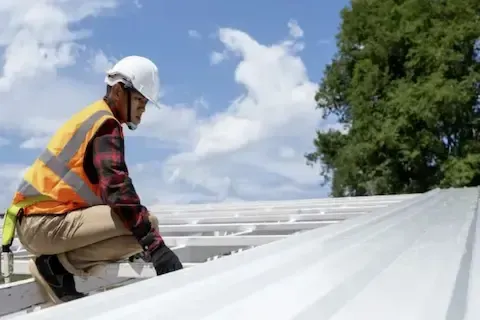 Commercial roofer kneeling to inspect metal panel seams on sun-lit rooftop