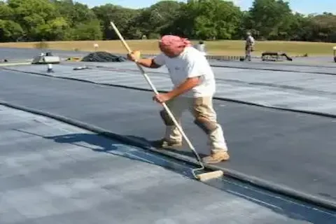 Worker spreading EPDM rubber roofing membrane across flat surface with long-handled roller