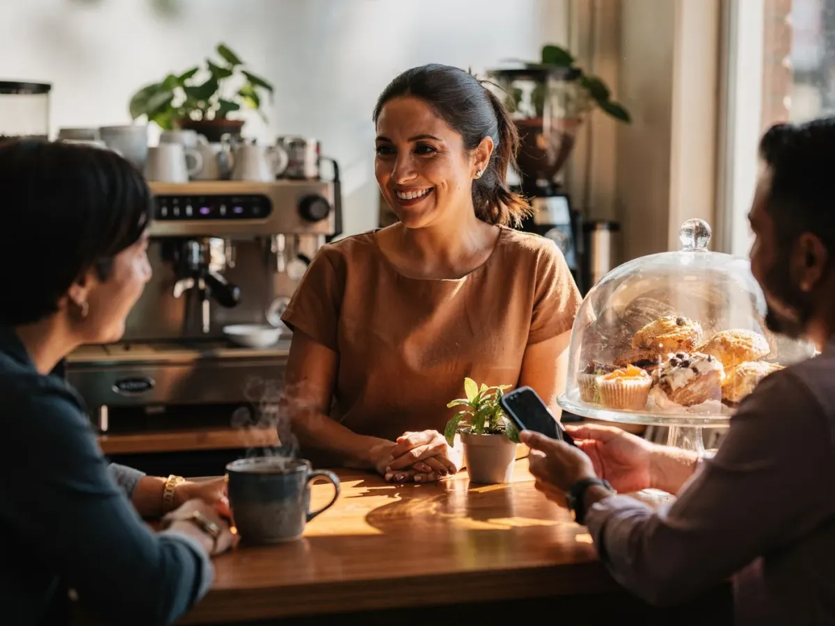Coffee shop owner smiling and chatting with two regular customers at her counter