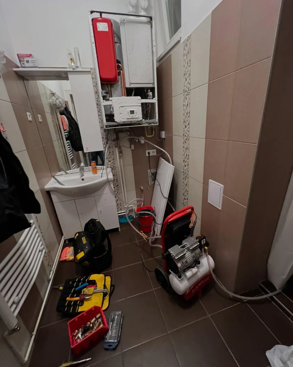 A small bathroom with brown tiles shows maintenance work in progress. Tools and a portable compressor are on the floor near a sink and wall-mounted boiler.