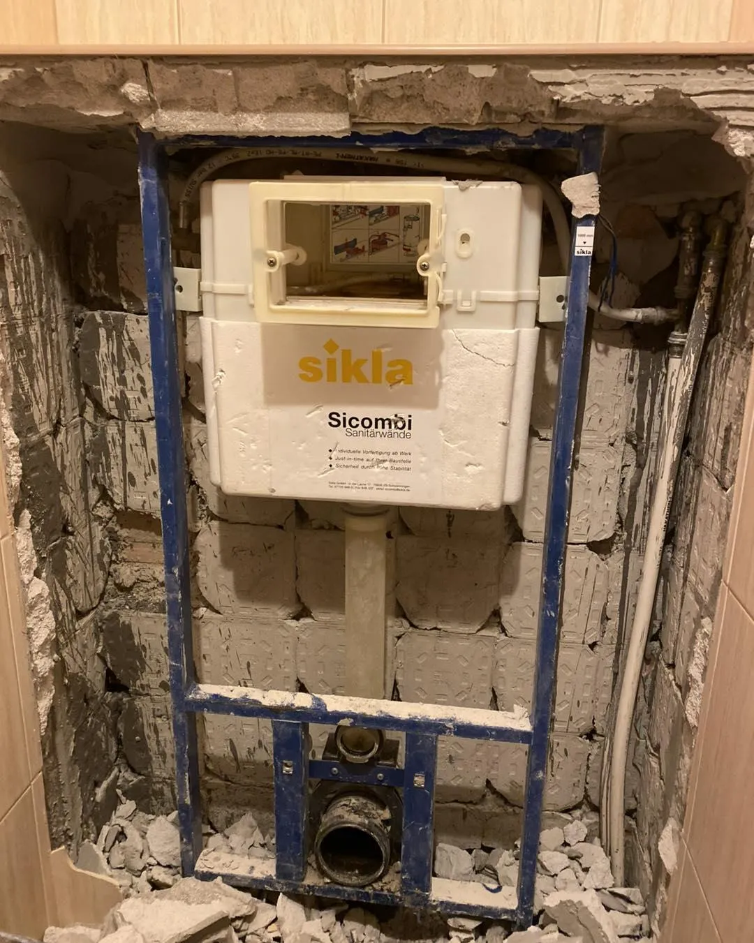 Exposed cistern installation in a bathroom wall. The wall is partially removed, revealing a white tank with visible pipes and debris around it.