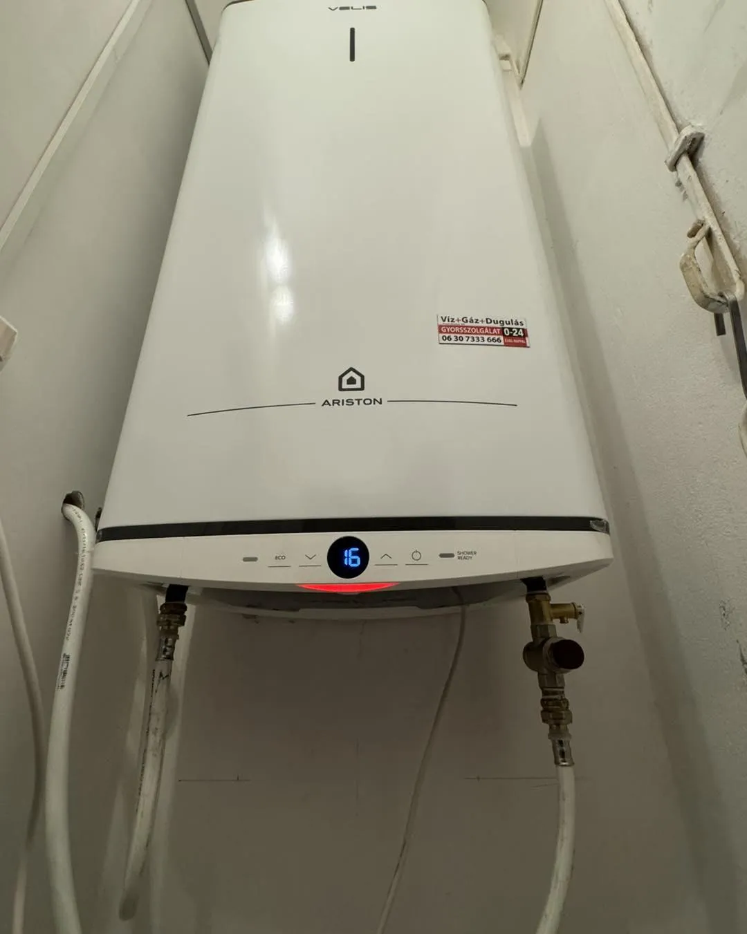 A wall-mounted white Ariston water heater in a narrow space, showing a digital display with the number 16. Pipes connect at the bottom.