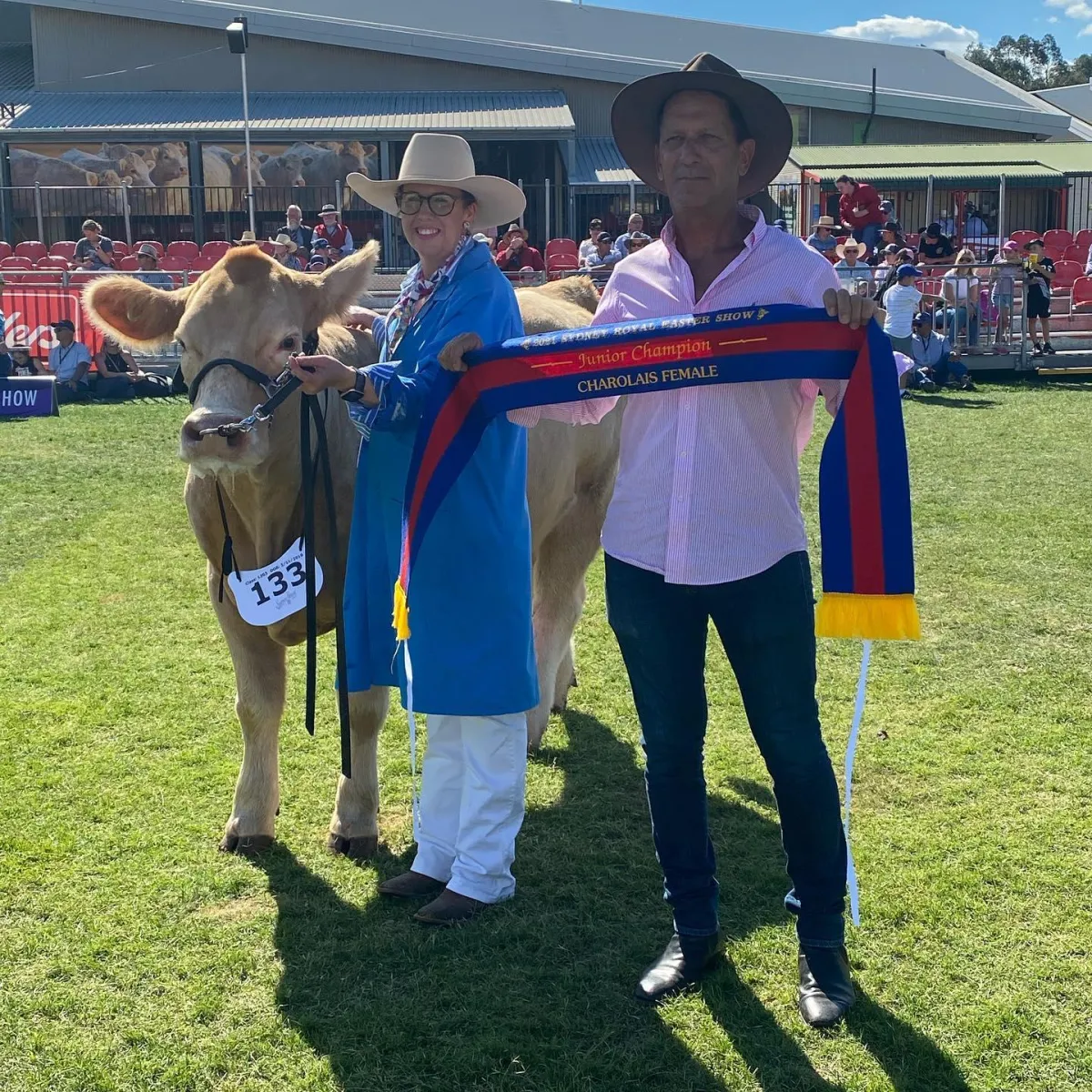 Greg Nicholson, owner of Black Duck Charolais, proudly displaying a 'Junior Champion Charolais Female' ribbon at a sunny outdoor cattle show, alongside a prize-winning Charolais heifer held by a smiling handler in blue.