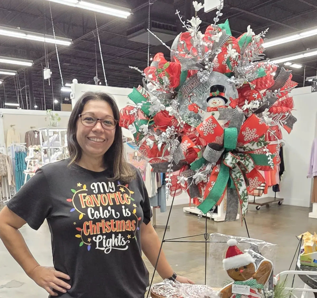 A smiling woman in her 30s, wearing a linen apron, holds a freshly crafted floral wreath in a sunlit modern studio. The background is softly blurred, with natural light and neutral tones, emphasizing the wreath's vibrant colors and the creator's pride.