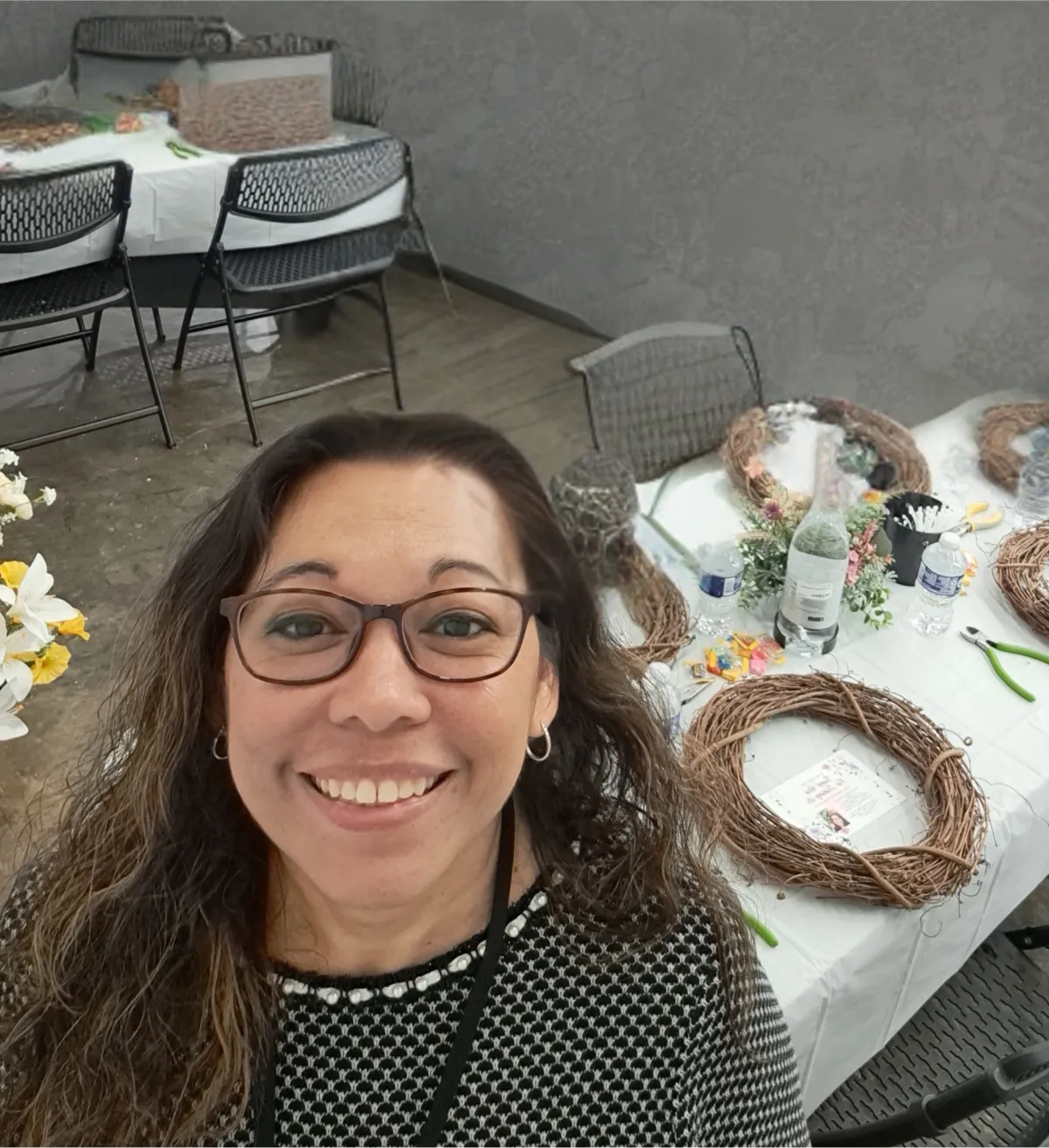Two diverse instructors, one middle-aged woman and one younger man, stand together at a worktable covered in wreath-making supplies. Both are smiling warmly, surrounded by greenery and ribbons, in a bright, welcoming craft studio.