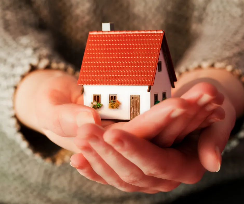 A person holds a small model house with a red roof and white walls in their cupped hands, symbolizing parents ready to sell home or connect with an as is home buyer.
