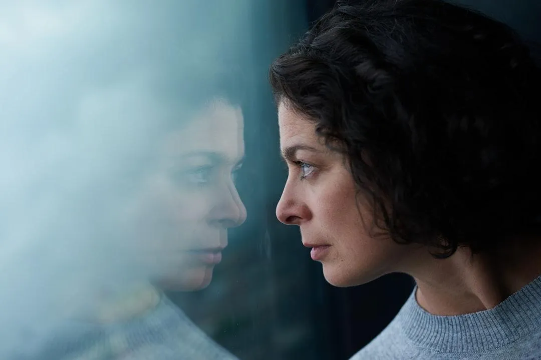 A woman with short, dark curly hair looks thoughtfully at her reflection in a window, wearing a light gray sweater—perhaps considering an as is home sale as an elder planning her next chapter.