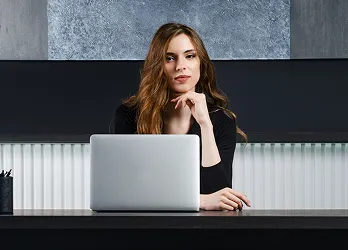 A woman with long wavy brown hair wearing a black top sits at a modern desk with a silver laptop. She rests her chin on her hand with a confident expression. The background features a dark and textured wall design.