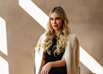 Professional woman with long blonde curls wearing a black outfit and a white coat, posing confidently in natural sunlight with a neutral-toned background.