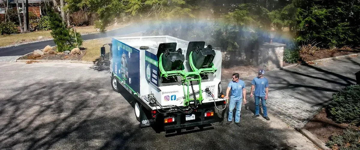 Clean Island Cans truck using hot water to sanitize garbage bins while two team members stand nearby under a visible rainbow mist.