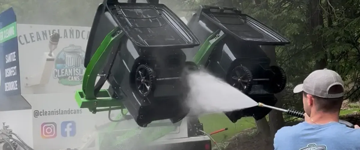 Worker using a high-pressure washer to clean upside-down trash bins on a cleaning truck