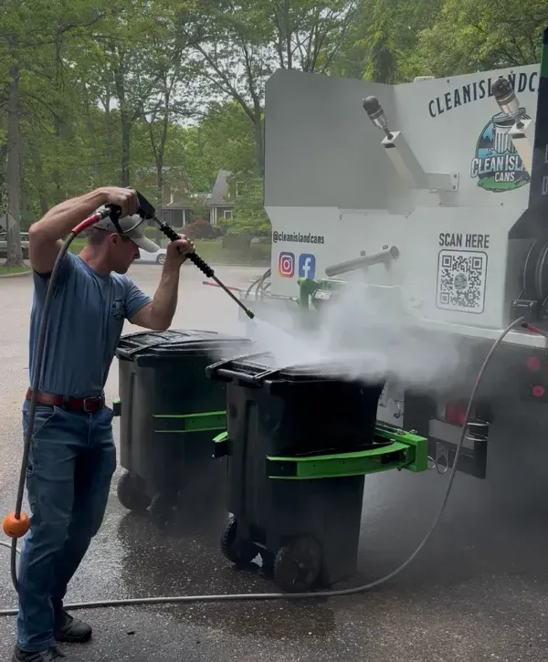 A Clean Island Cans technician power-washing residential trash bins beside a bin-cleaning truck, with steam rising during the cleaning process.