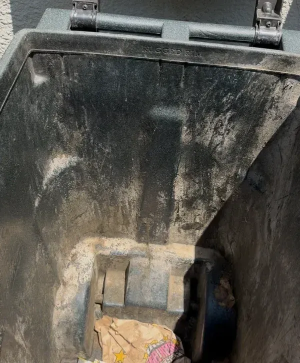 A close-up view looking down into a dark, dirty trash can with a small amount of crumpled trash at the bottom.