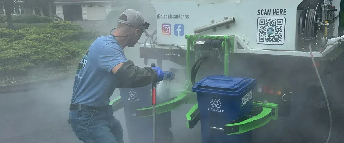 A man in a baseball cap and goggles power washes a blue recycling bin attached to a commercial cleaning truck.