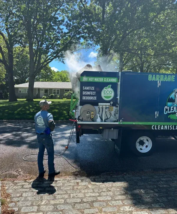 A man in a cap and t-shirt operating a hot water cleaning system from a branded truck on a sunny street.