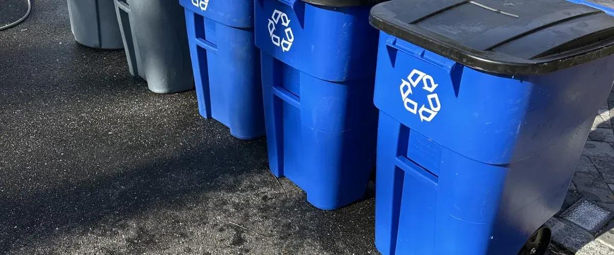 Several blue recycling bins with white recycle symbols are neatly lined up on a speckled asphalt surface.