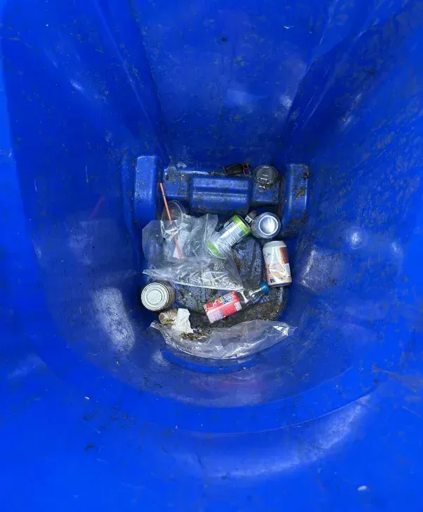 A top-down view inside a dirty blue recycling bin filled with various cans, plastic bags, and discarded items.