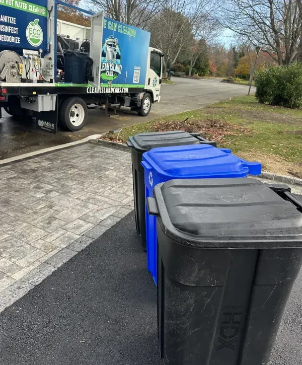 A blue and white garbage can cleaning truck parked on an asphalt driveway next to three outdoor trash cans.