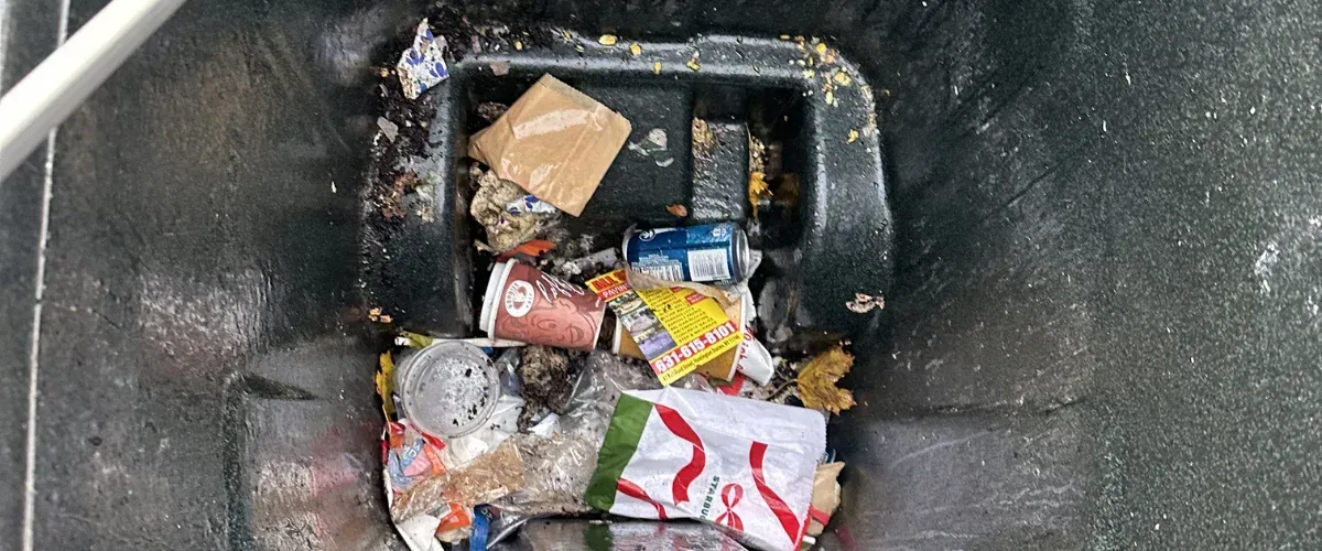 A top-down view inside a dirty blue recycling bin filled with various cans, plastic bags, and discarded items.