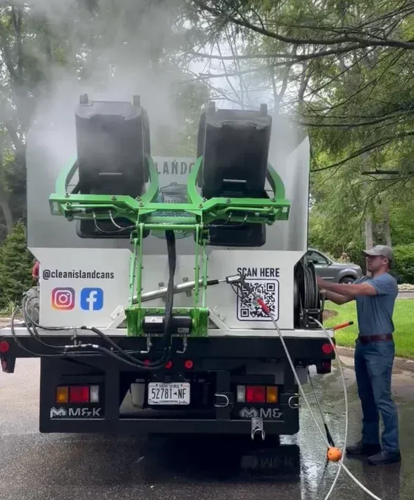 A man in a cap power washes two large black trash cans, tilted on a white truck with green accents.