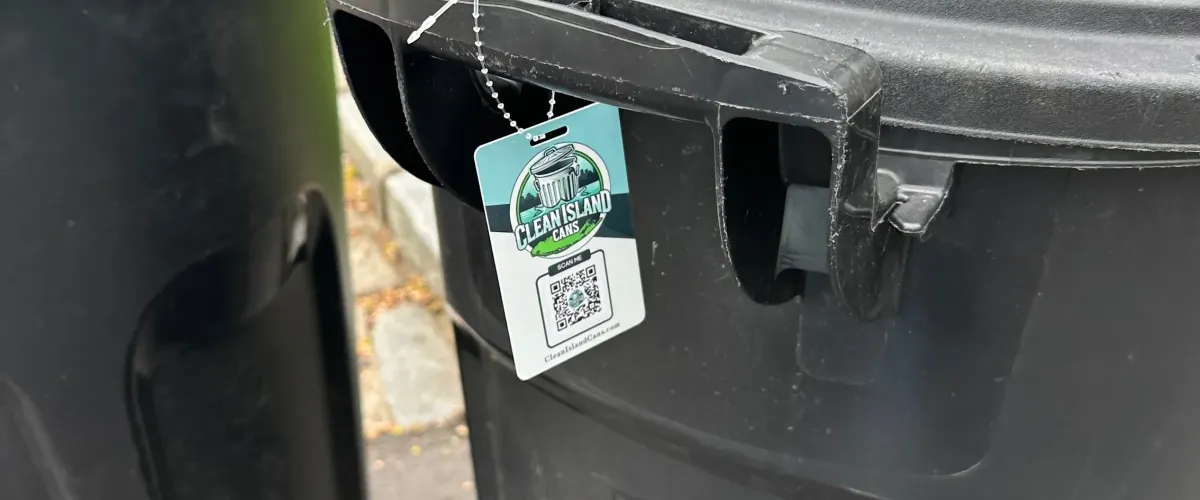 A close-up of a 'Clean Island Cans' tag with a QR code hanging from the handle of a black trash can.
