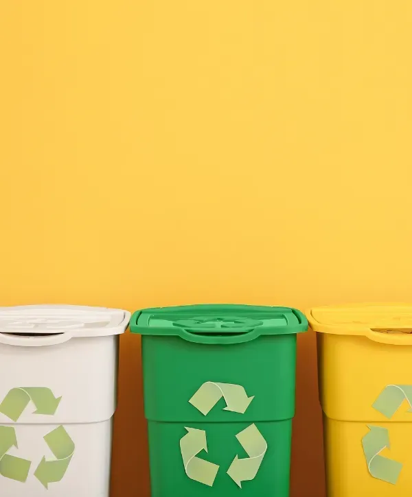 White, green, and yellow recycling bins with recycle symbols placed against a bright yellow wall.
