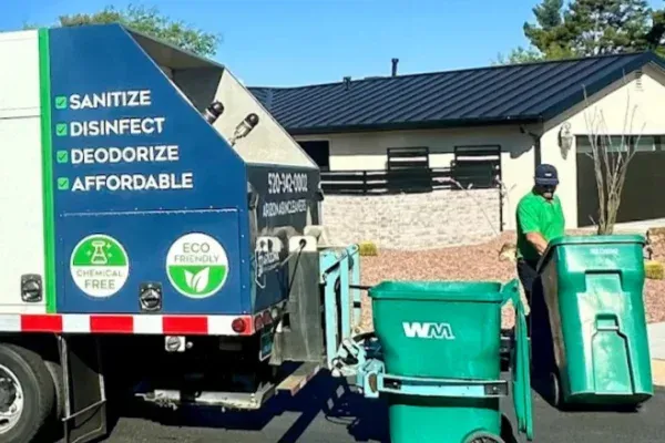 A sanitation worker in a green shirt and hat loads a green WM trash bin into a waste management truck.