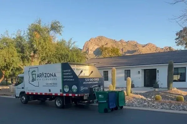 An Arizona Bin Cleaners truck with three trash cans on the side of a street in front of a white house with mountains in the background.