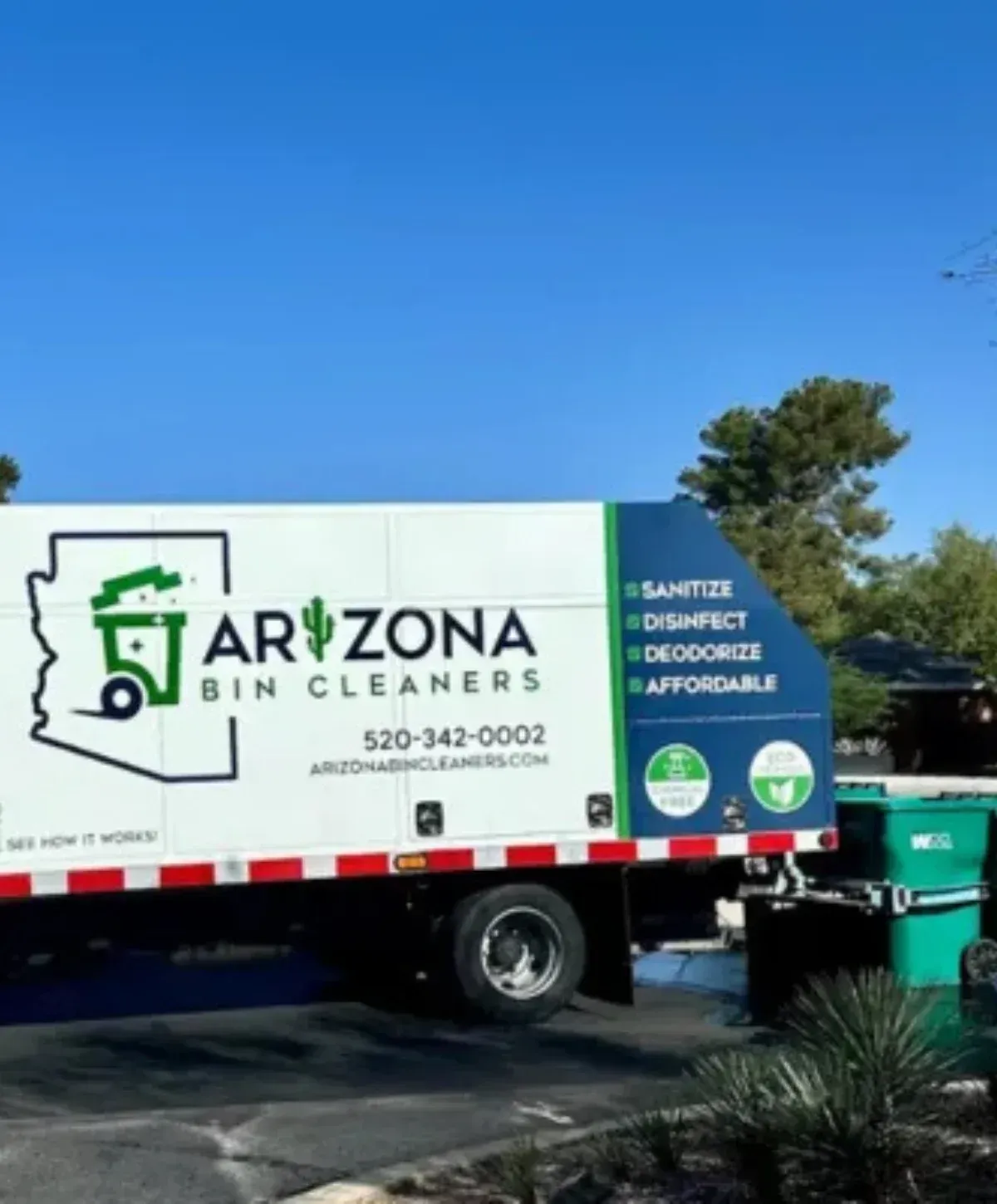 A white Arizona Bin Cleaners truck cleans two waste bins in front of a modern white desert home with cacti and mountains.
