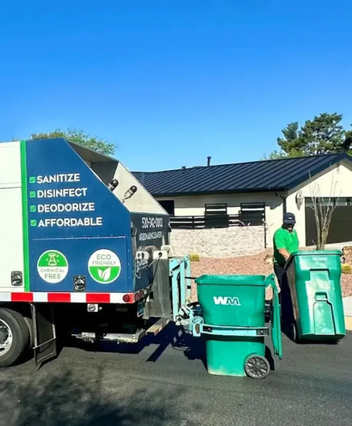 A worker in a green shirt cleans a green waste bin beside a white and blue sanitation truck on a sunny day.