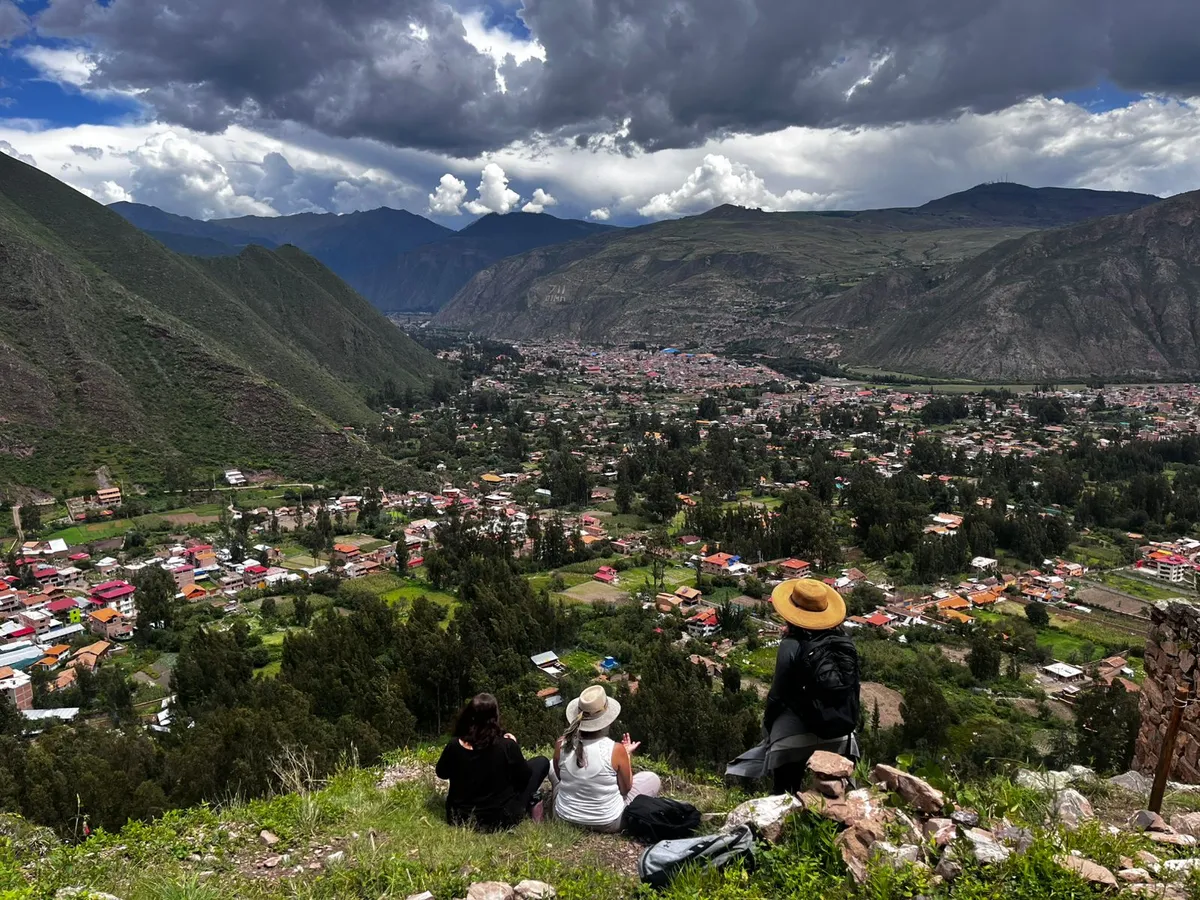 Three women sit on a hillside above Urubamba, looking out over the Sacred Valley beneath storm clouds